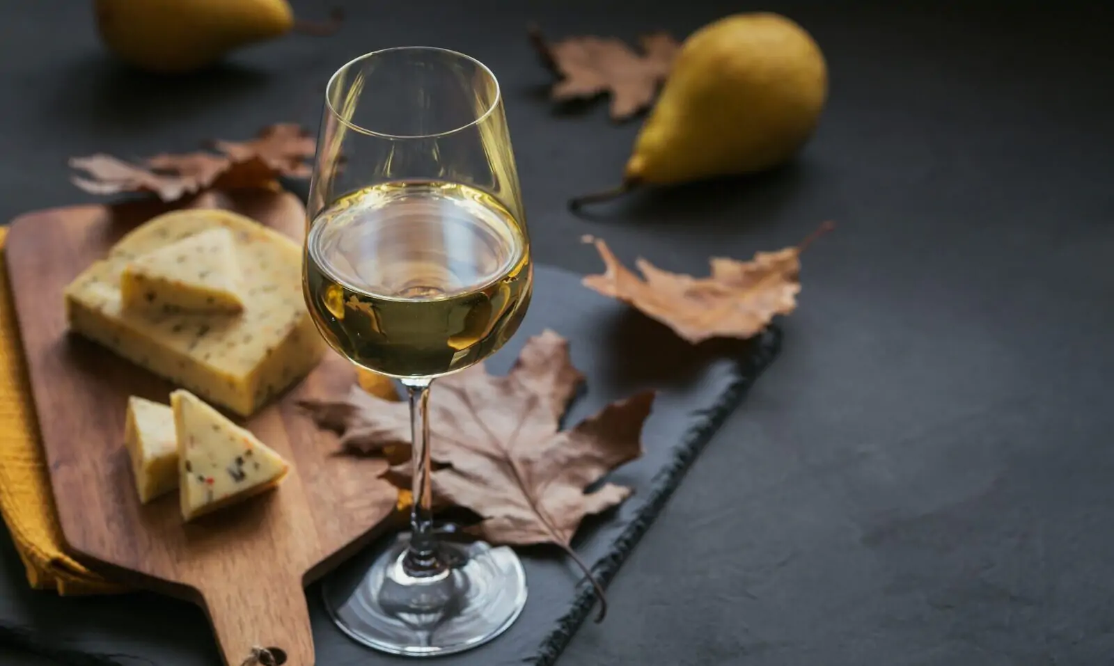 A glass of white wine served with cheese in a cutting board on dark background